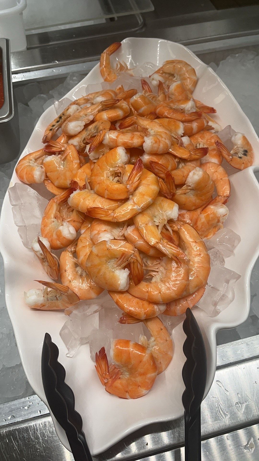 Cooked shrimp on ice, served in a white scalloped dish, with serving tongs.