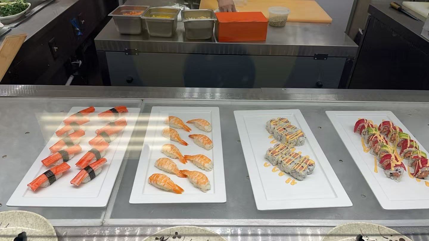 Sushi display: plates of nigiri and rolls behind glass counter.