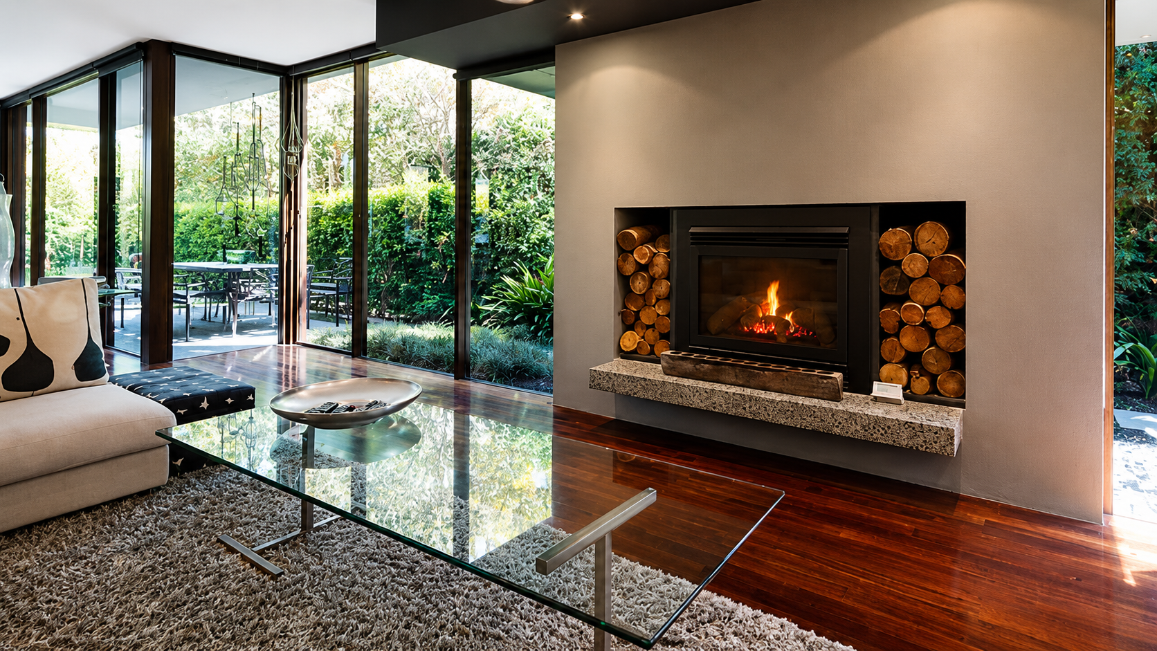 Modern living room with glass walls, a fireplace, stone accent wall, and view of a lush garden
