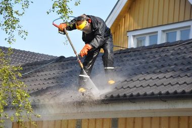 Worker using a pressure washer on a house roof, spraying water and debris in daylight