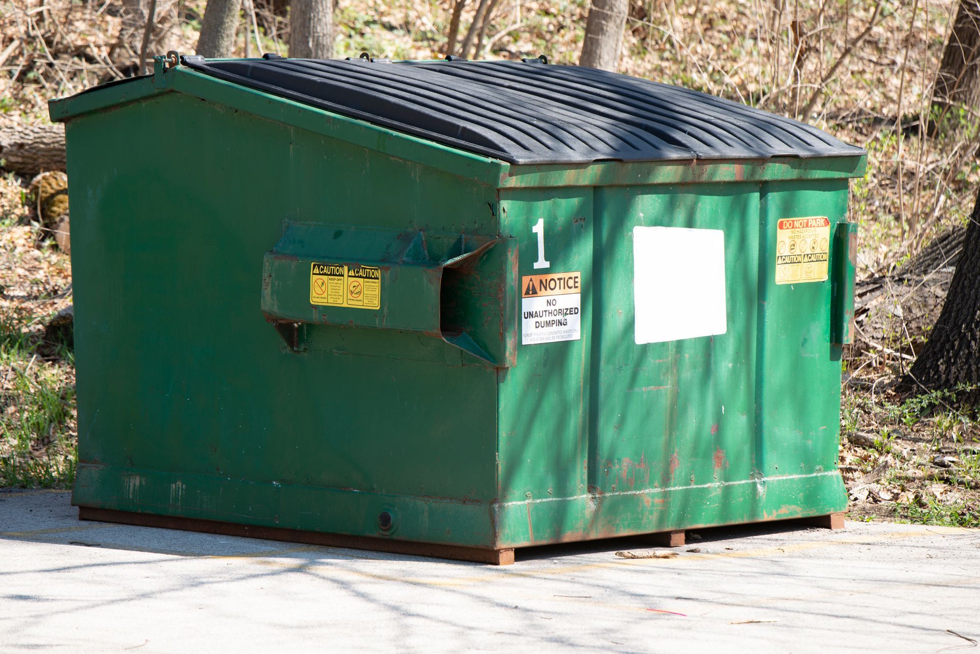 Green dumpster with black lid and warning labels on a paved area outdoors