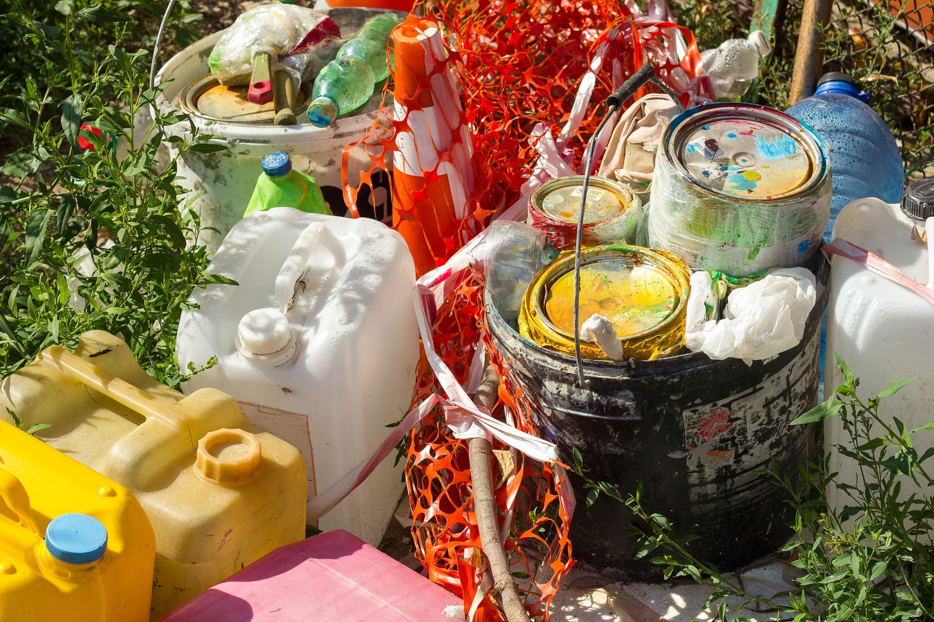 Assorted plastic jugs and paint cans piled outdoors amid greenery and orange safety netting