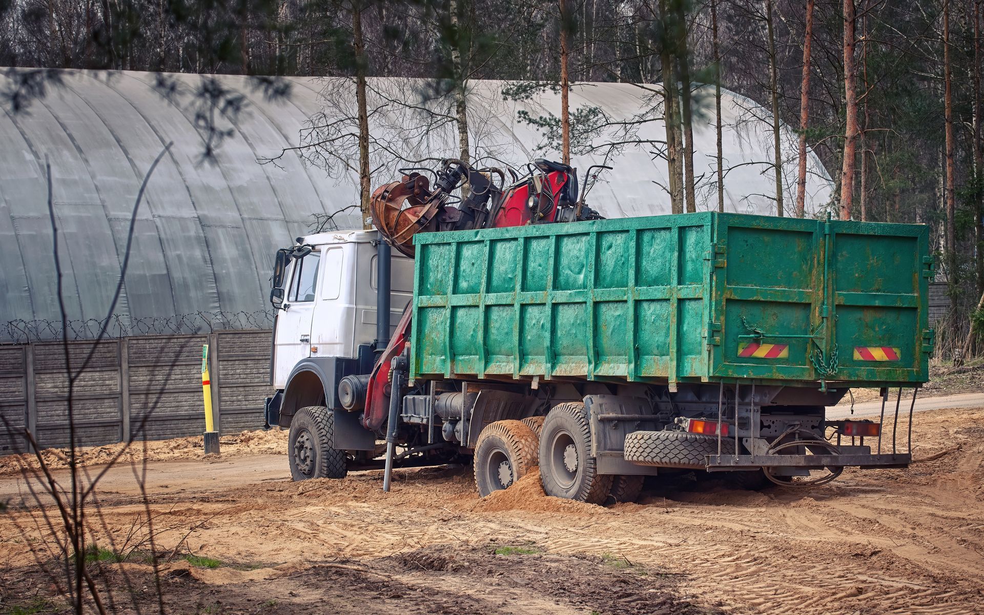 White truck with green cargo bed parked in a muddy wooded area near a greenhouse structure