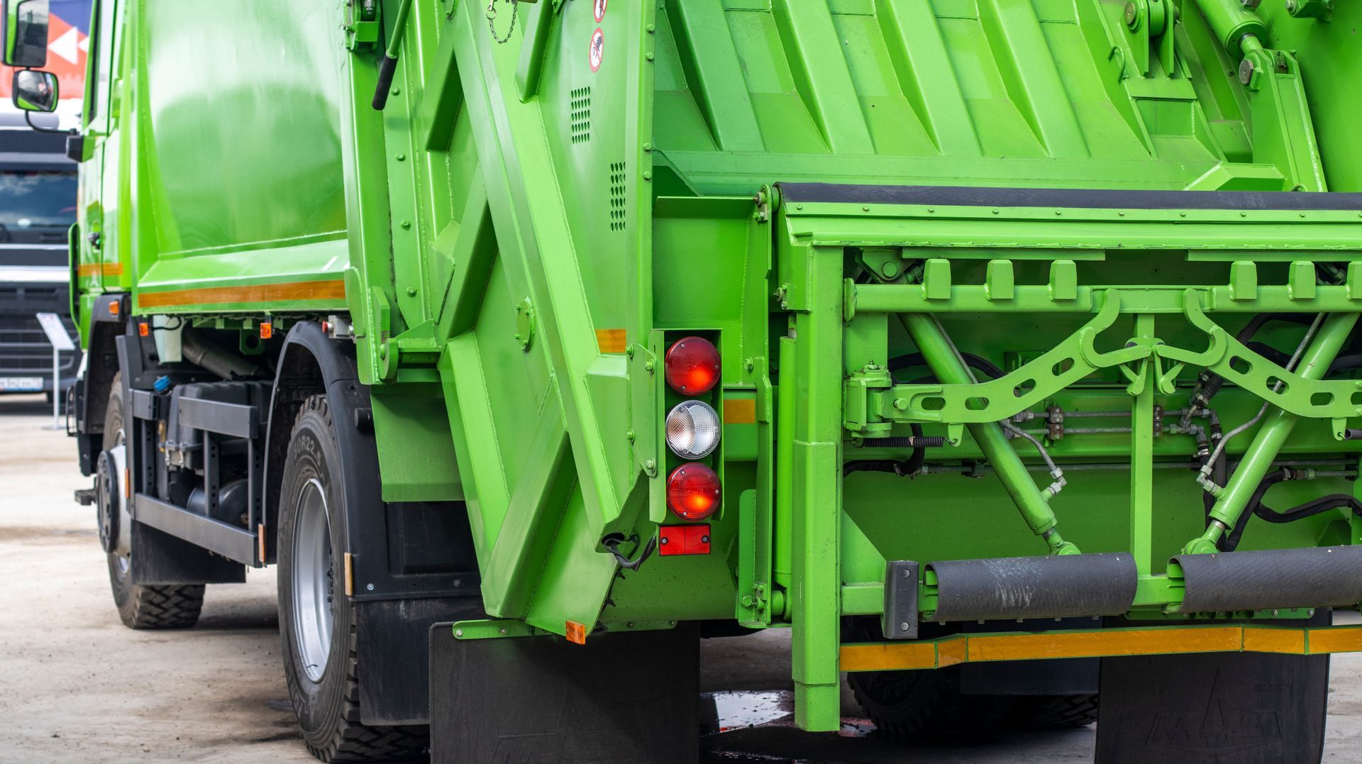 Bright green garbage truck rear view on a street, with hydraulic lifting mechanism visible.
