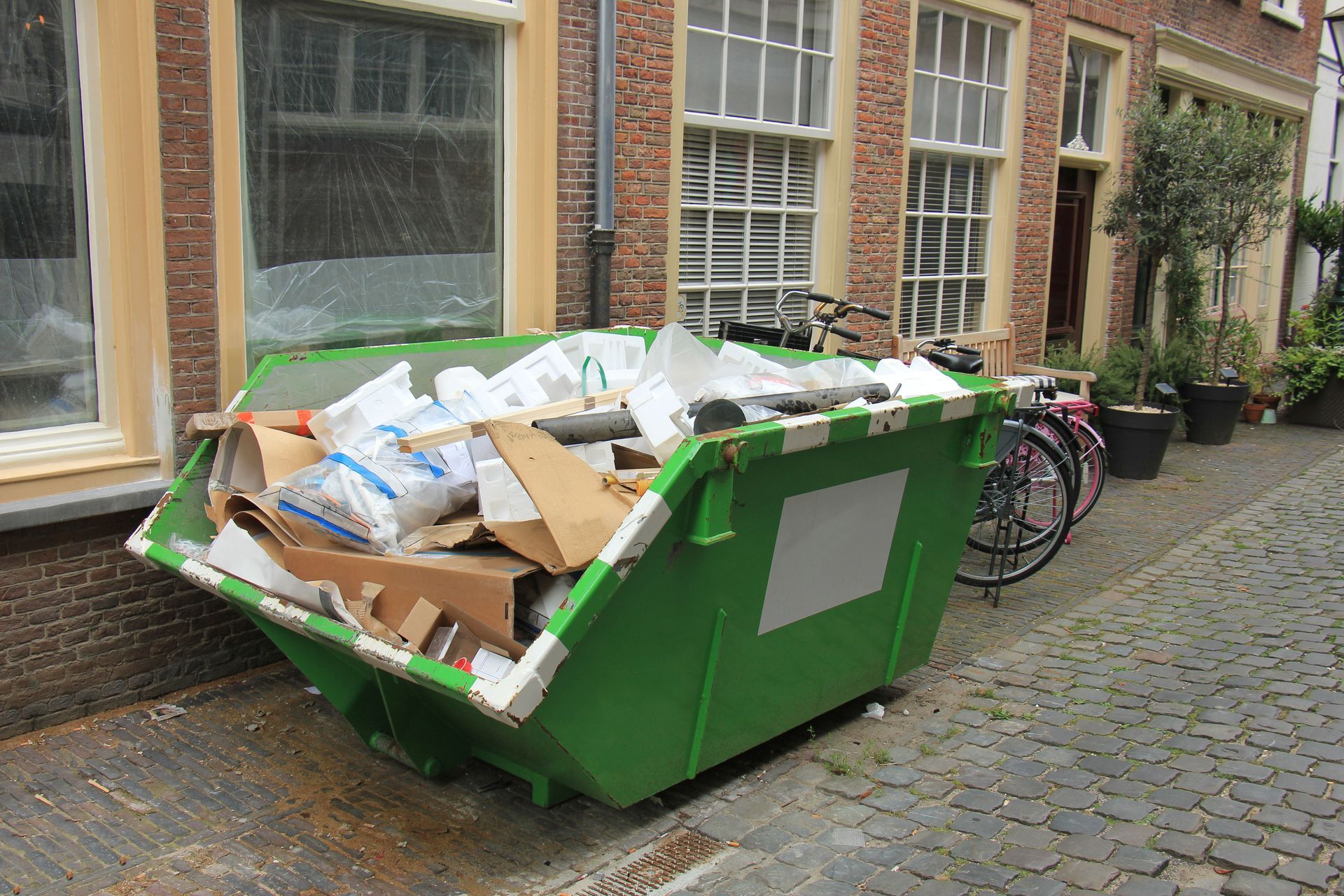 Green dumpster overflowing with cardboard and trash on a cobblestone street beside brick buildings and bicycles