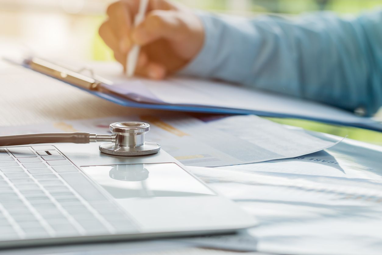 A doctor is writing on a clipboard next to a laptop and a stethoscope.