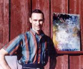 Man in striped shirt stands by colorful abstract painting against red wood siding.