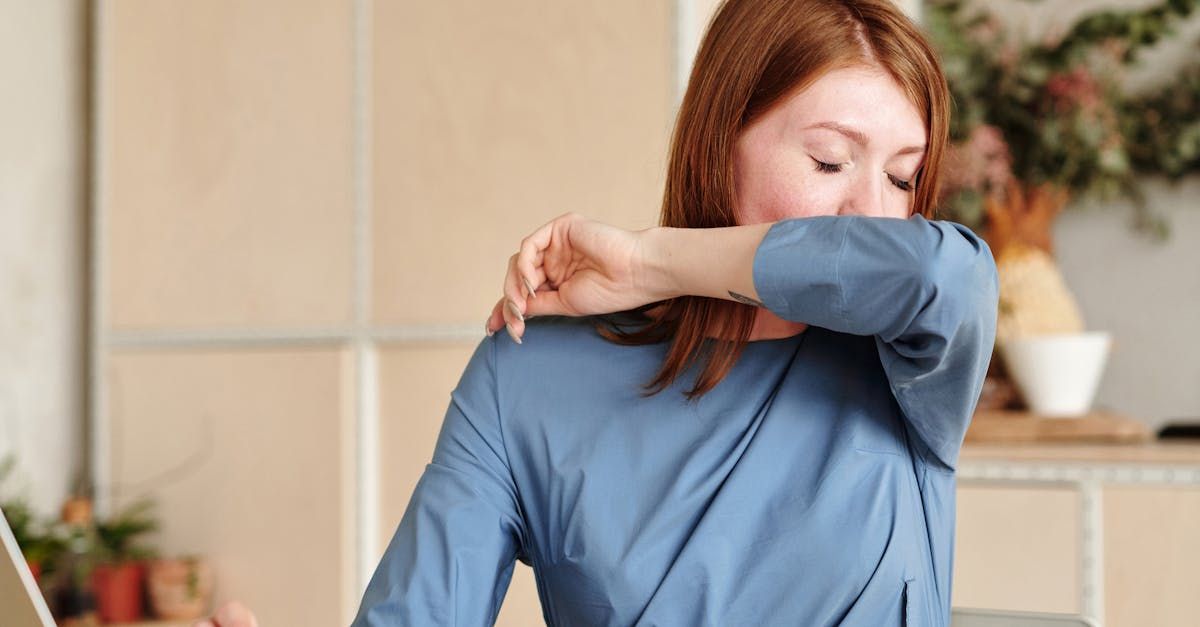 A woman is sitting at a desk with a laptop and coughing into her elbow.