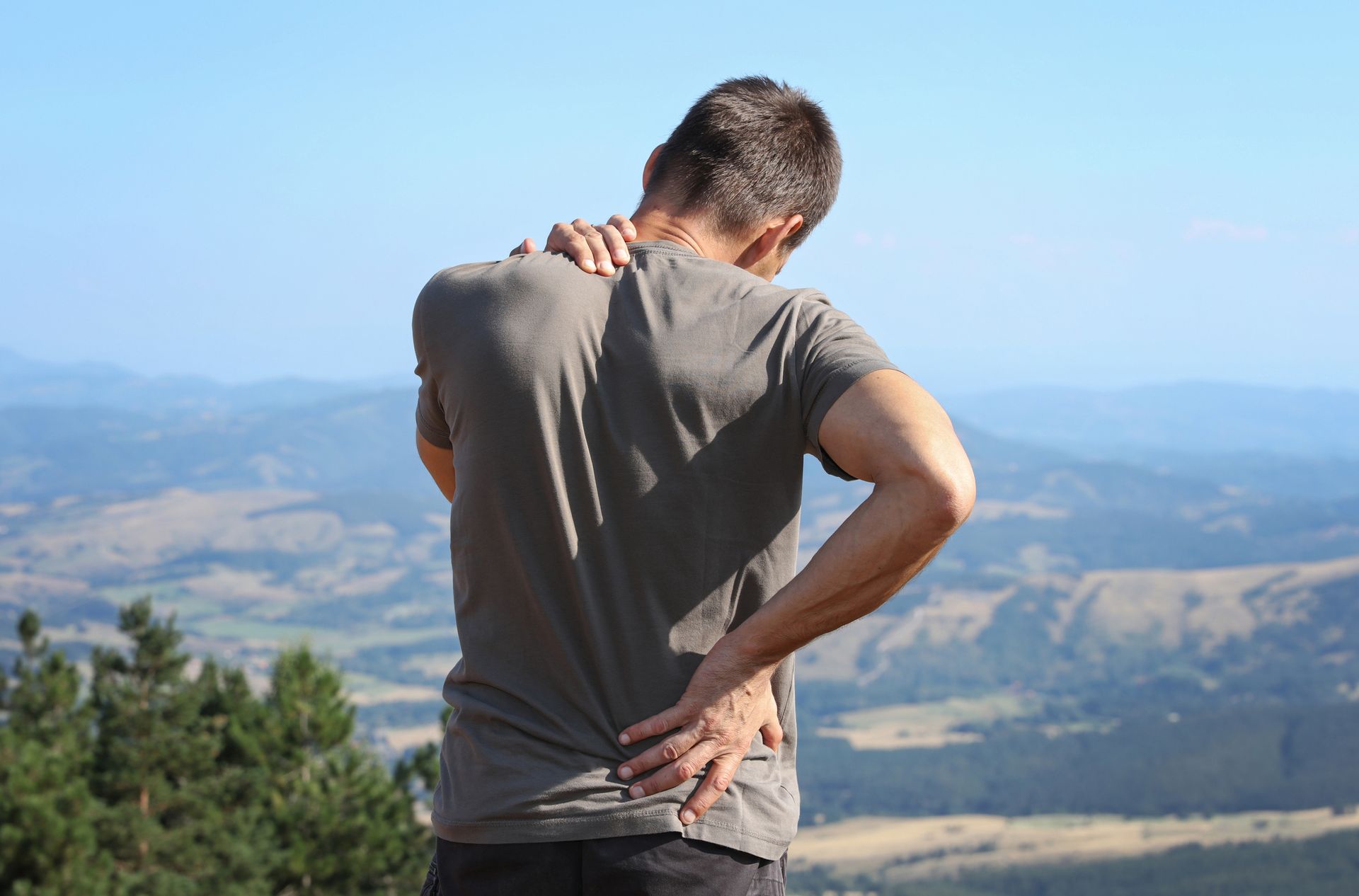 Man outdoors, holding back and neck, suggesting pain, with scenic background.