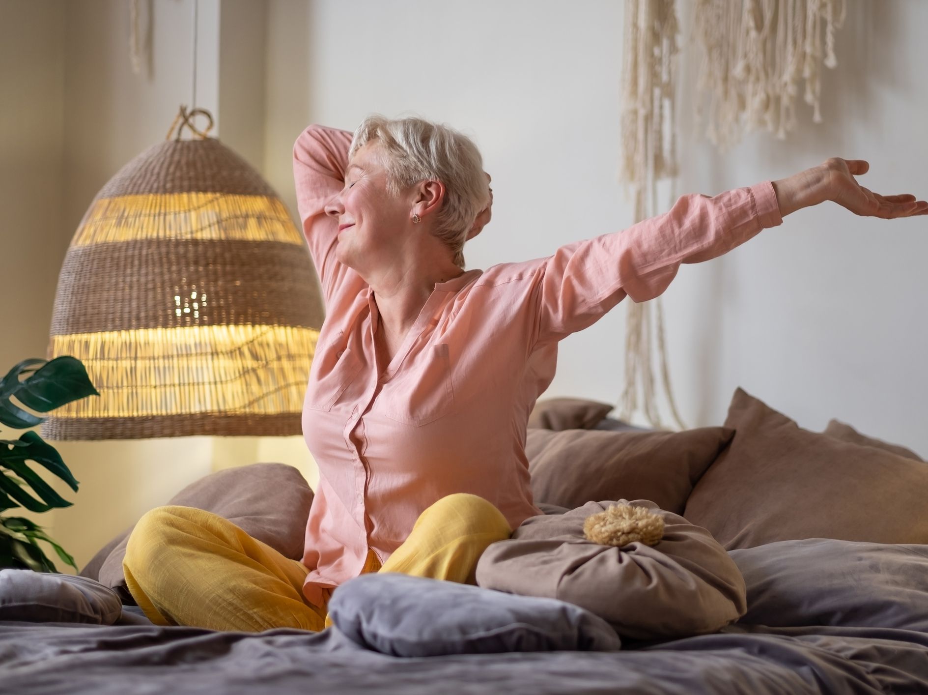 Woman with short blonde hair stretches on a bed, smiling, in a soft-lit bedroom.