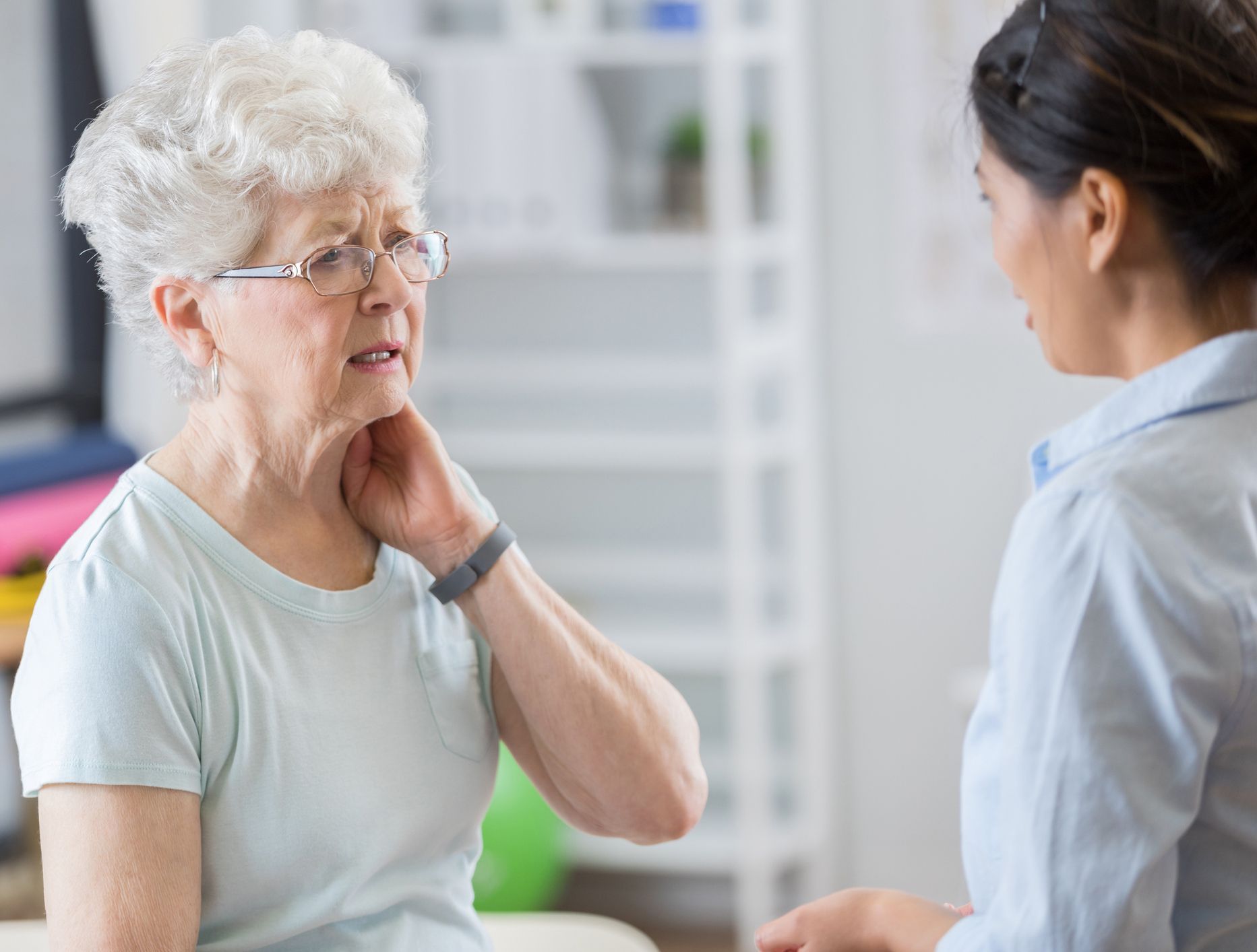 An elderly woman holds her neck, talking to a healthcare worker in a medical setting.
