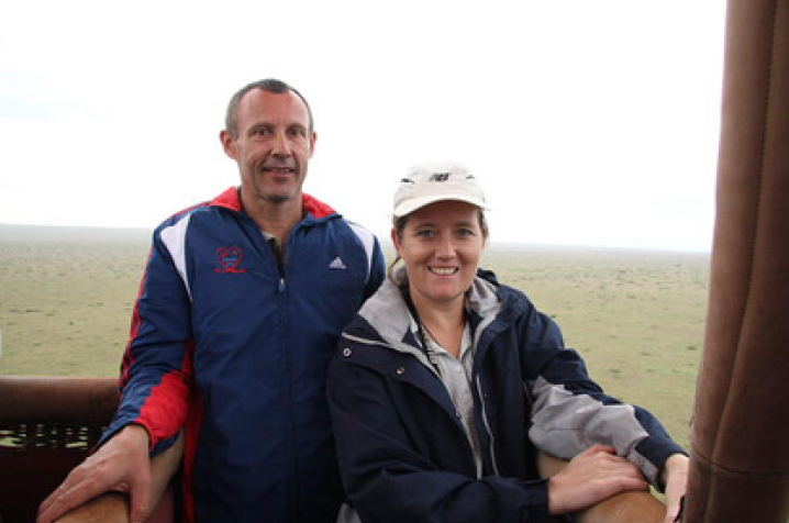 A man and a woman smile, posing inside a hot air balloon over a vast, flat grassland.