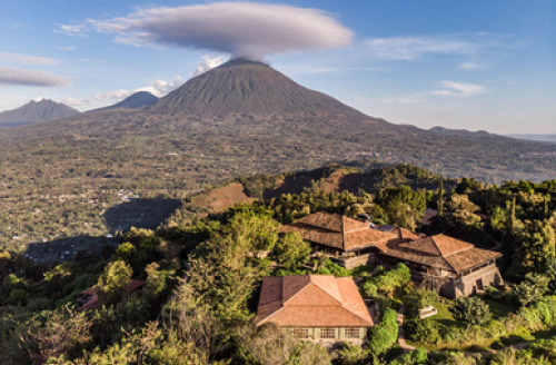 Aerial view of a lodge with orange tile roofs nestled in lush greenery, with a large volcano and cloud in the background.