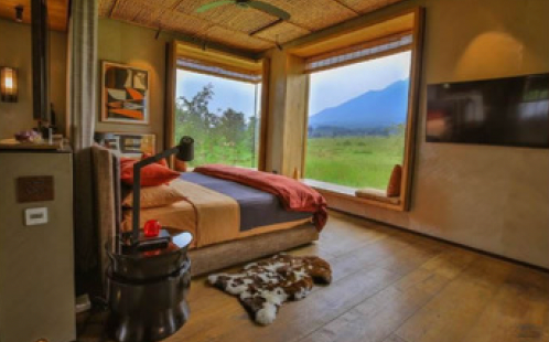 Bedroom with large windows offering a view of a green landscape and a mountain. Interior features a bed, TV, and a small decorative rug.