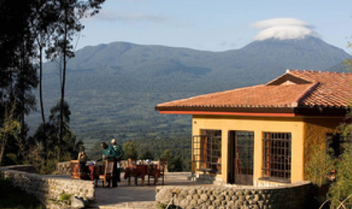 Terrace of a yellow building with red-tiled roof, a mountain with a cloud on top in the background. People dine at a table.