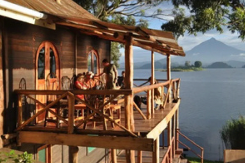 Wooden cabin with a deck overlooking a lake and mountain. People are seated on the deck.