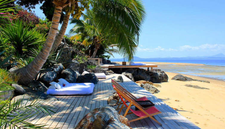 Beach scene with wooden deck, lounge beds, palm trees, and ocean view.