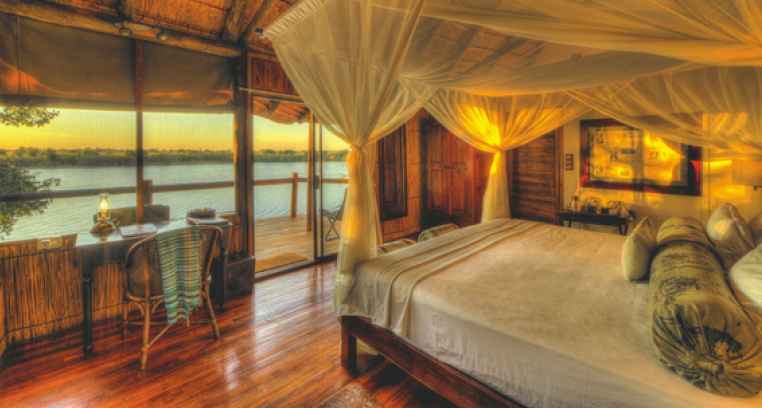Bedroom with a bed draped in white fabric, overlooking a river at sunset. Wooden walls, floors, and a desk with a view.