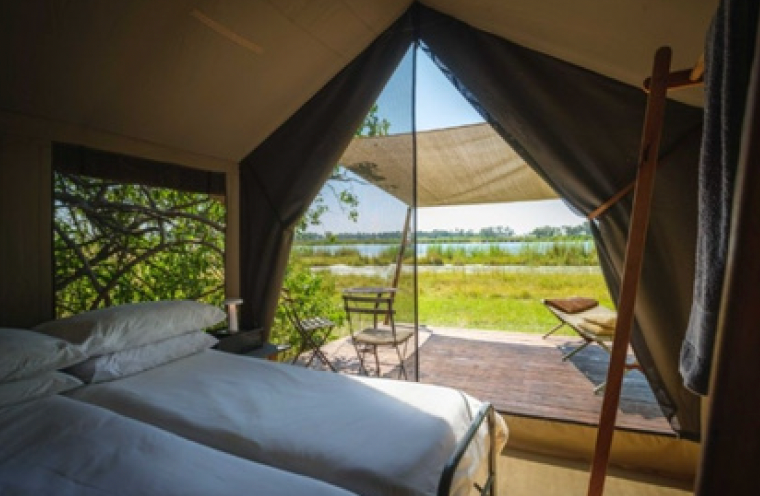 Interior of a canvas tent with two white-sheeted beds, open doorway looking out onto a lake and surrounding vegetation.