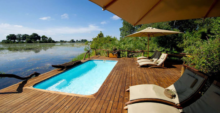 Swimming pool on a wooden deck overlooking a river, with lounge chairs and an umbrella. Lush green trees and blue sky in the background.