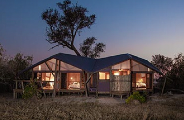 Safari lodge at dusk, showcasing lit interiors and dark blue roof. The wooden structure is set in a grassy field with trees.