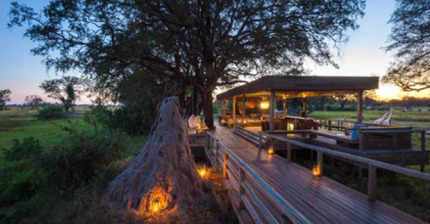 Wooden walkway lit by lanterns leading to a covered outdoor structure at dusk, in a grassy African landscape.