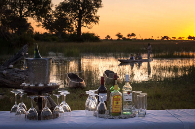 Sunset scene with a lakeside bar set up with drinks and glasses, canoe in the background.