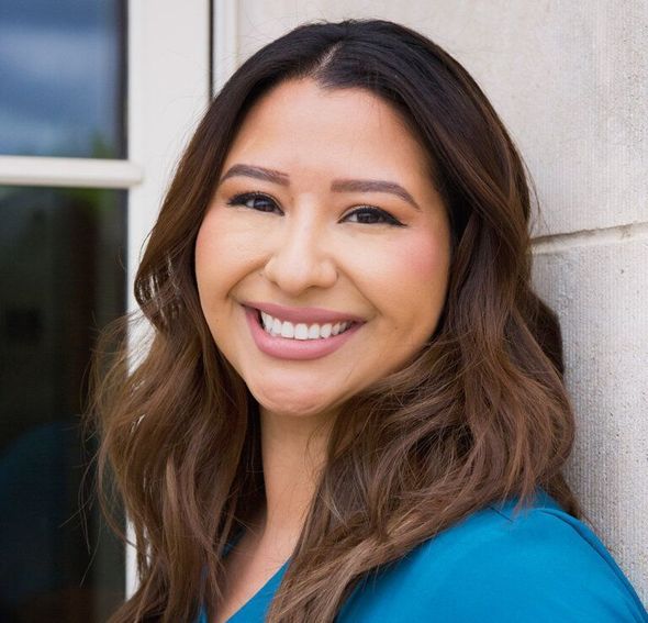 A woman in a blue shirt is smiling and leaning against a wall.