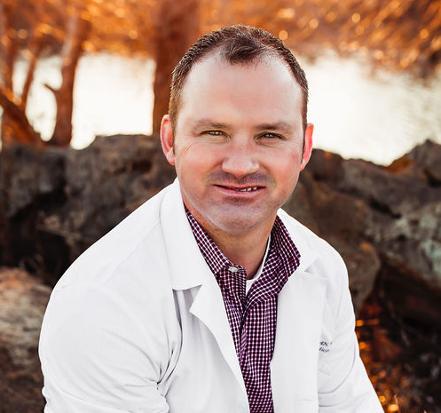 A man in a white lab coat is sitting on a rock looking at the camera.