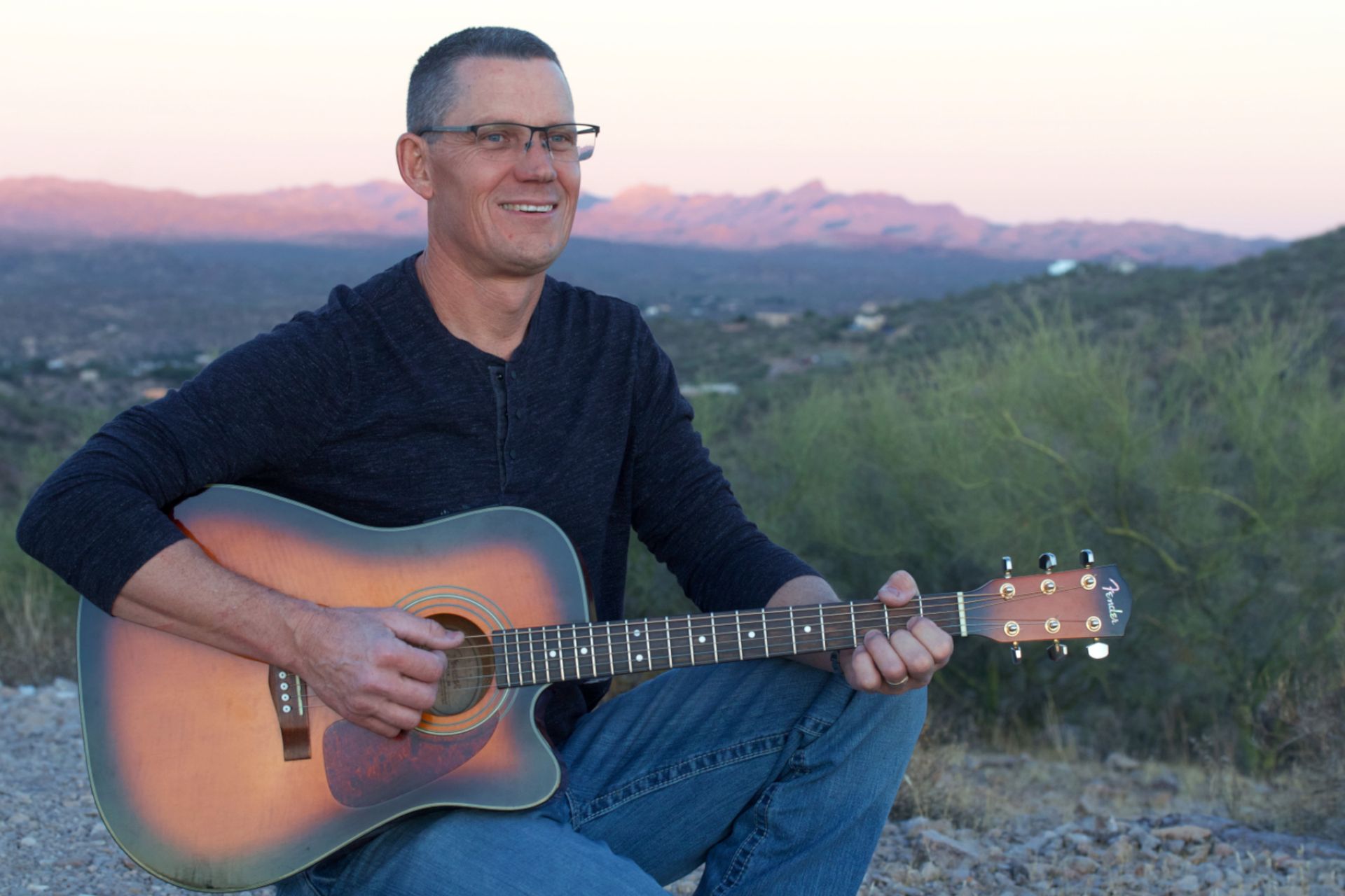 A man is kneeling down while playing an acoustic guitar.