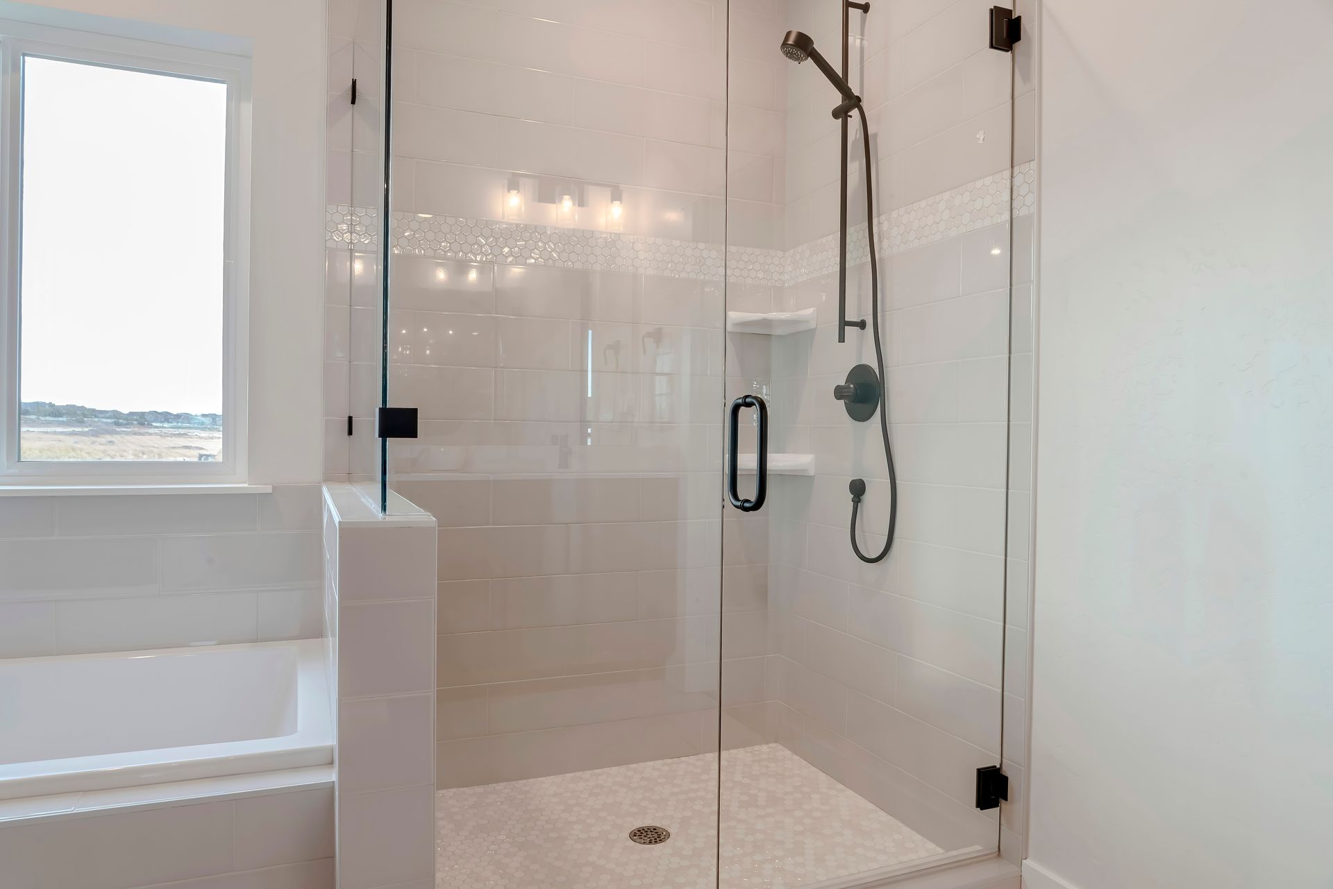 A modern shower with a clear glass door, white tile, and black fixtures. A window is to the left.
