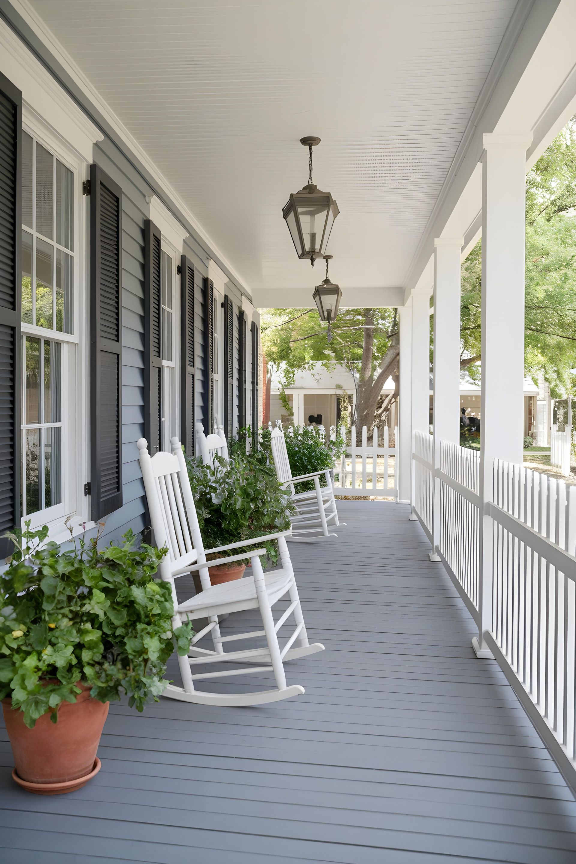 A covered porch with white rocking chairs, plants, and lanterns. The porch is painted grey.