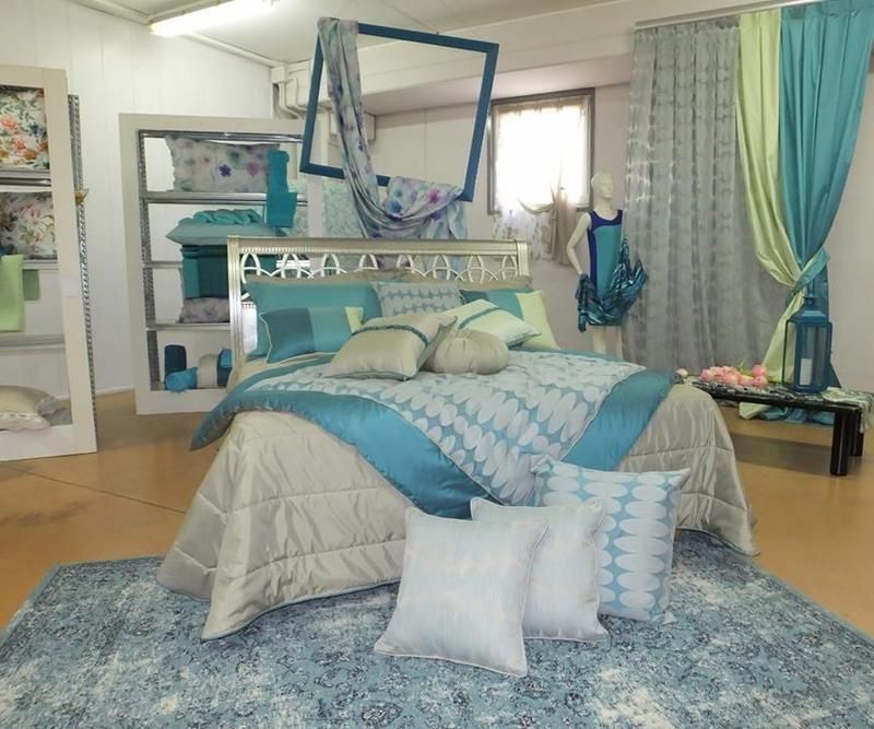 Esposizione della camera da letto nei toni del turchese e del grigio, con letto, tappeto, tende e dettagli decorativi.