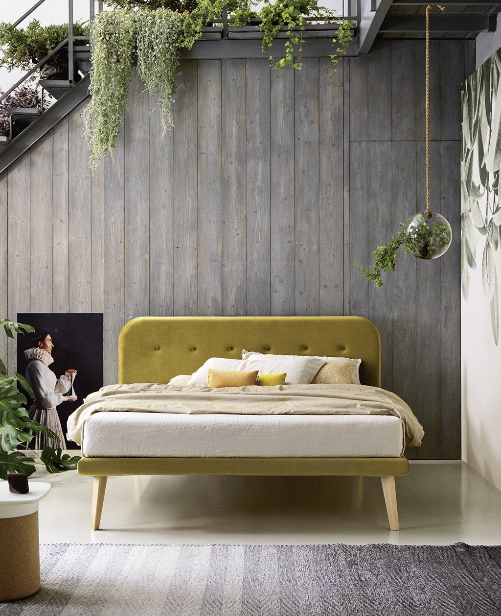 Letto in una camera moderna con parete in cemento e piante sospese. Testiera e gambe verde oliva.