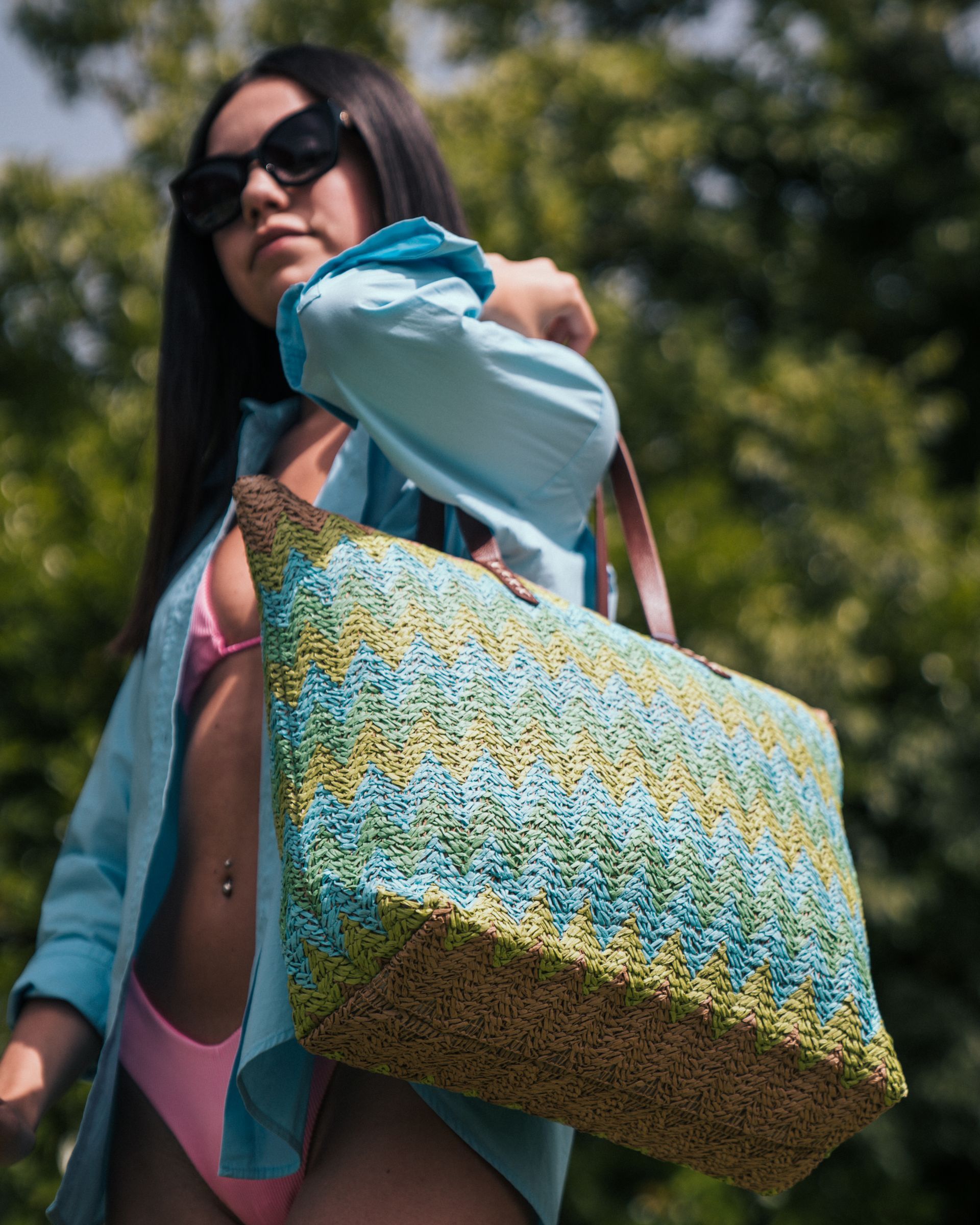 Una donna con occhiali da sole e camicia blu porta una borsa di tela intrecciata. Indossa un bikini rosa su uno sfondo verde.