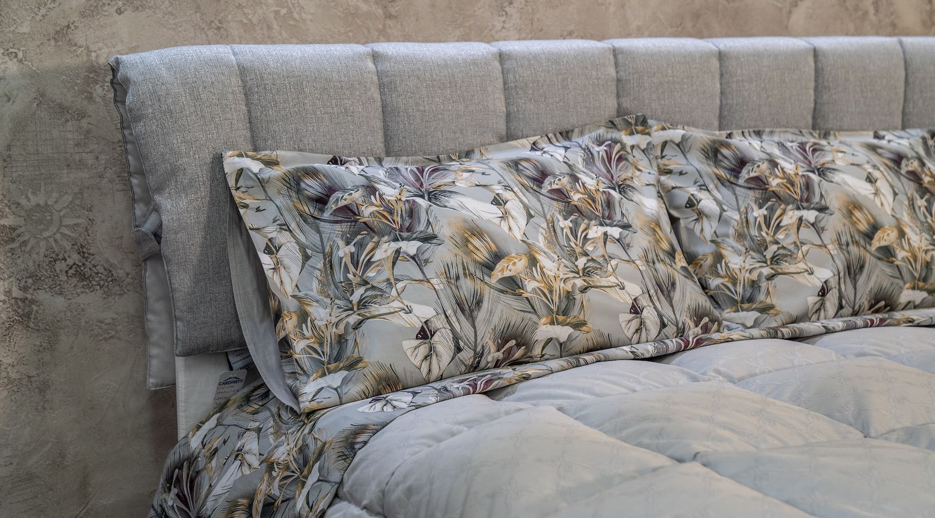 Letto con testiera imbottita grigia, cuscini floreali e piumone grigio.