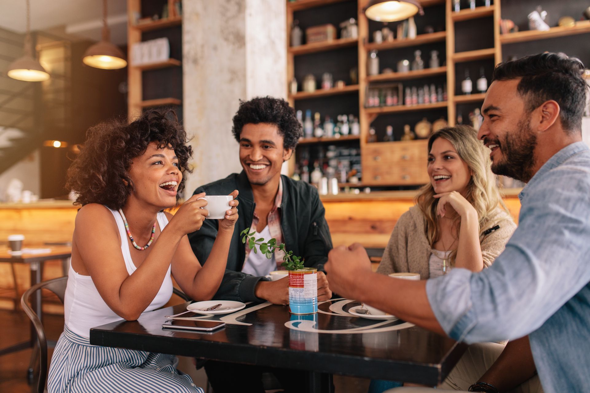 Four people laughing together at a cafe table, drinks present.