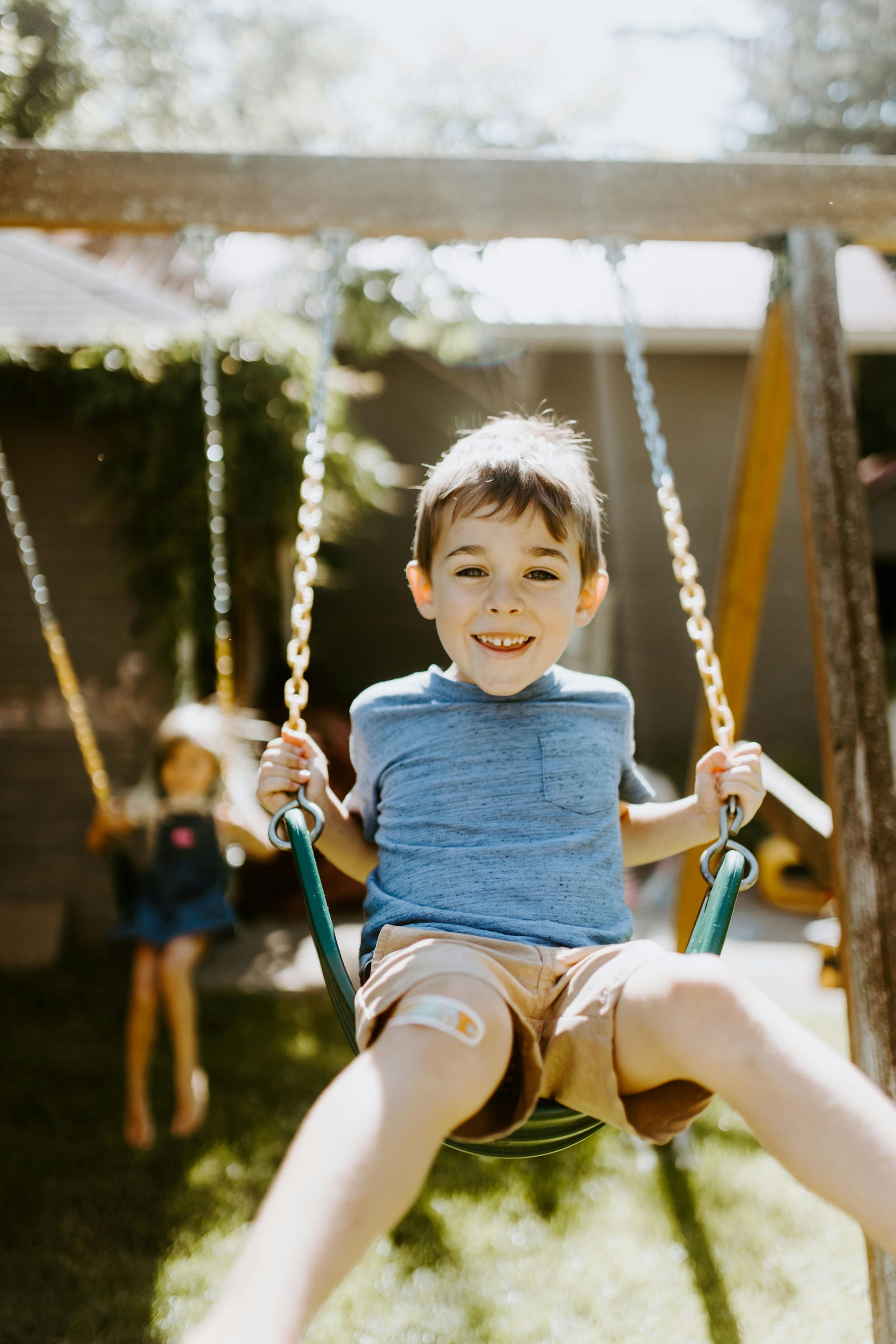 Boy smiling on a swing set, bandage on leg. Another child swings in the background, backyard setting.