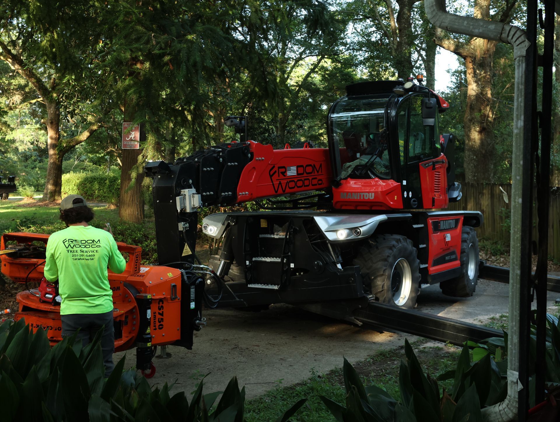 Man operating orange & black forestry machine, parked on gravel in a yard.