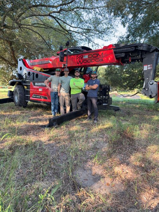 Four men pose in front of a red construction machine in a grassy area under trees.