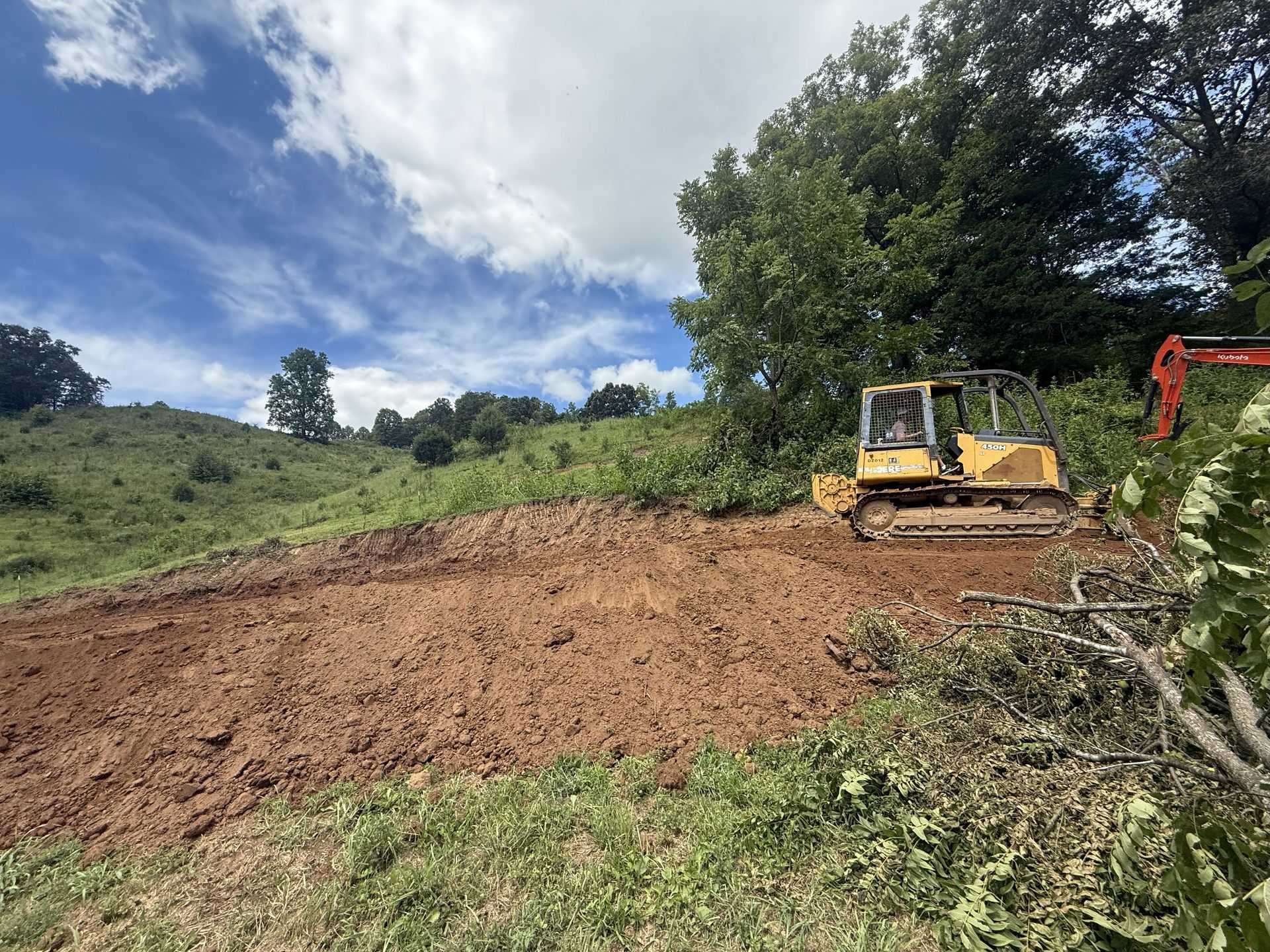 A bulldozer clears earth on a hillside. An excavator and trees are visible in the background.