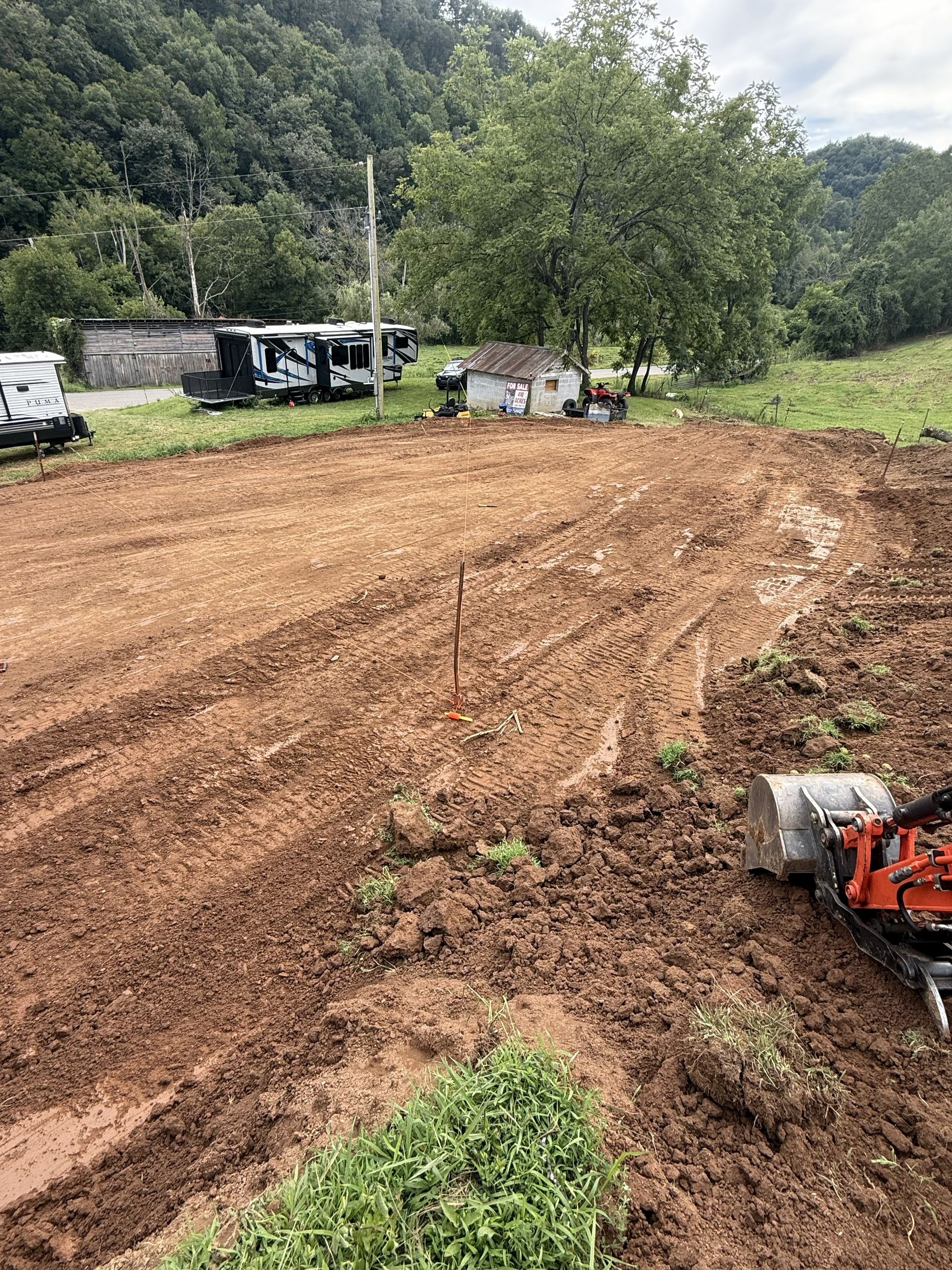 Brown dirt field, recently tilled, with small building and RV in the background, near a forested hill.