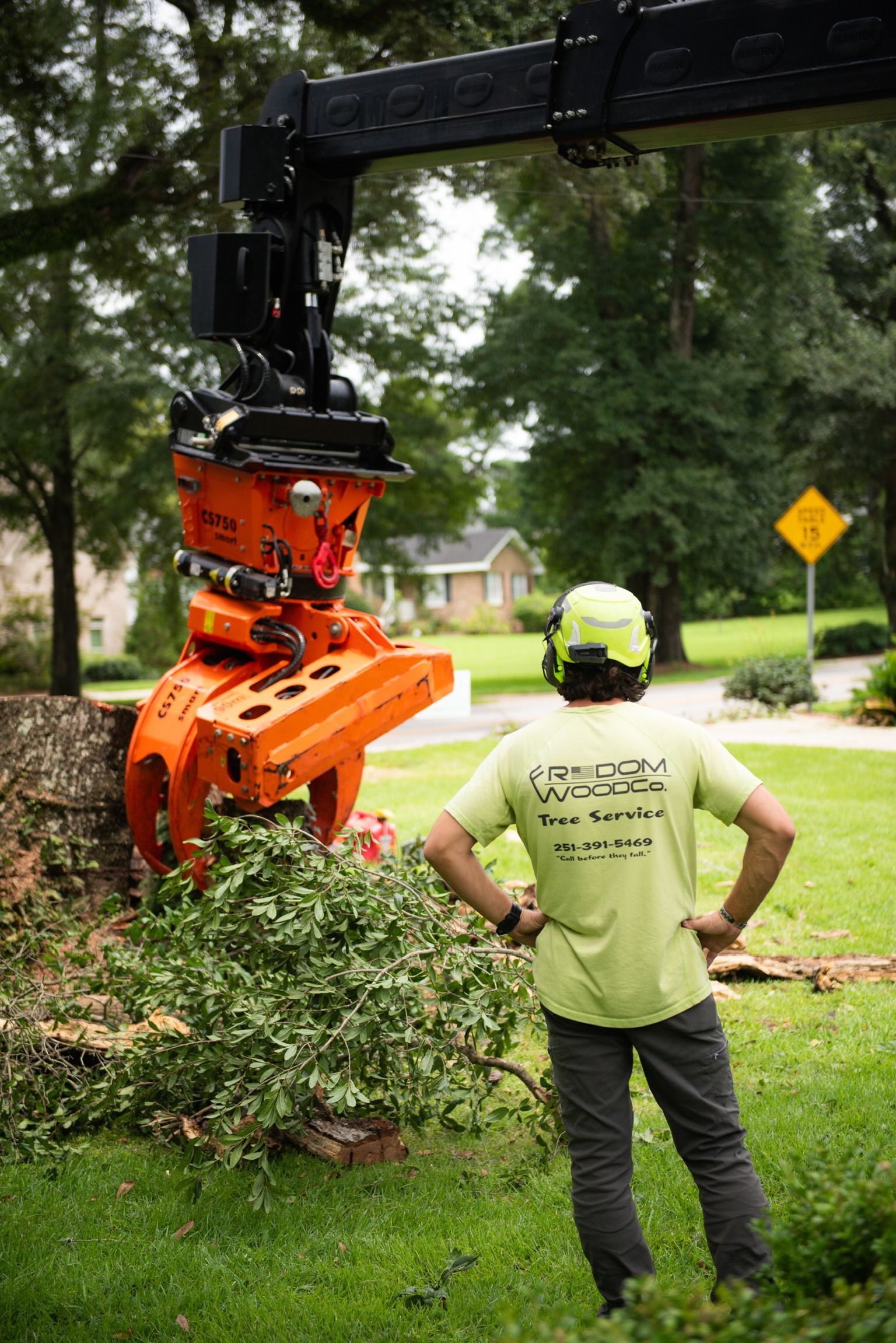 A tree service worker watches an orange mechanical tree cutter at work; grassy yard setting.