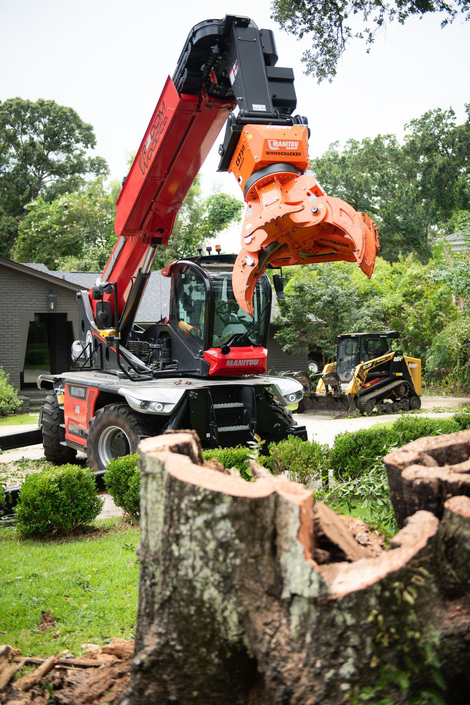 Red telehandler with grapple arm over a tree stump in front of a building.