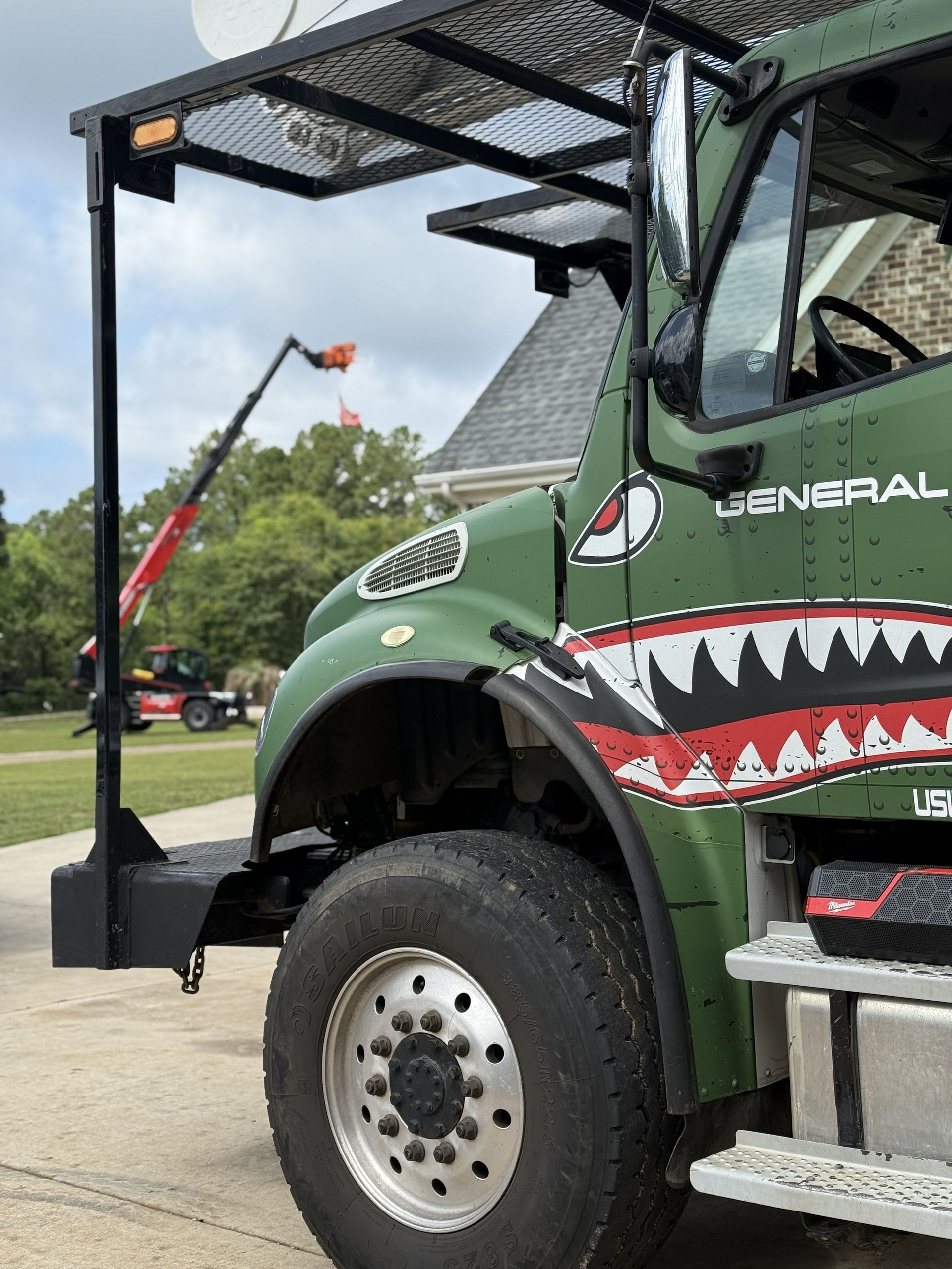 Green truck with shark mouth art, black rack, and large tires parked outside a house.
