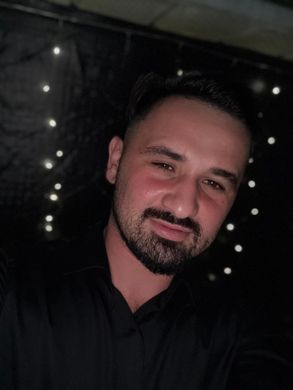 Man with a beard, smiling, wearing a black shirt, with small lights in the background.