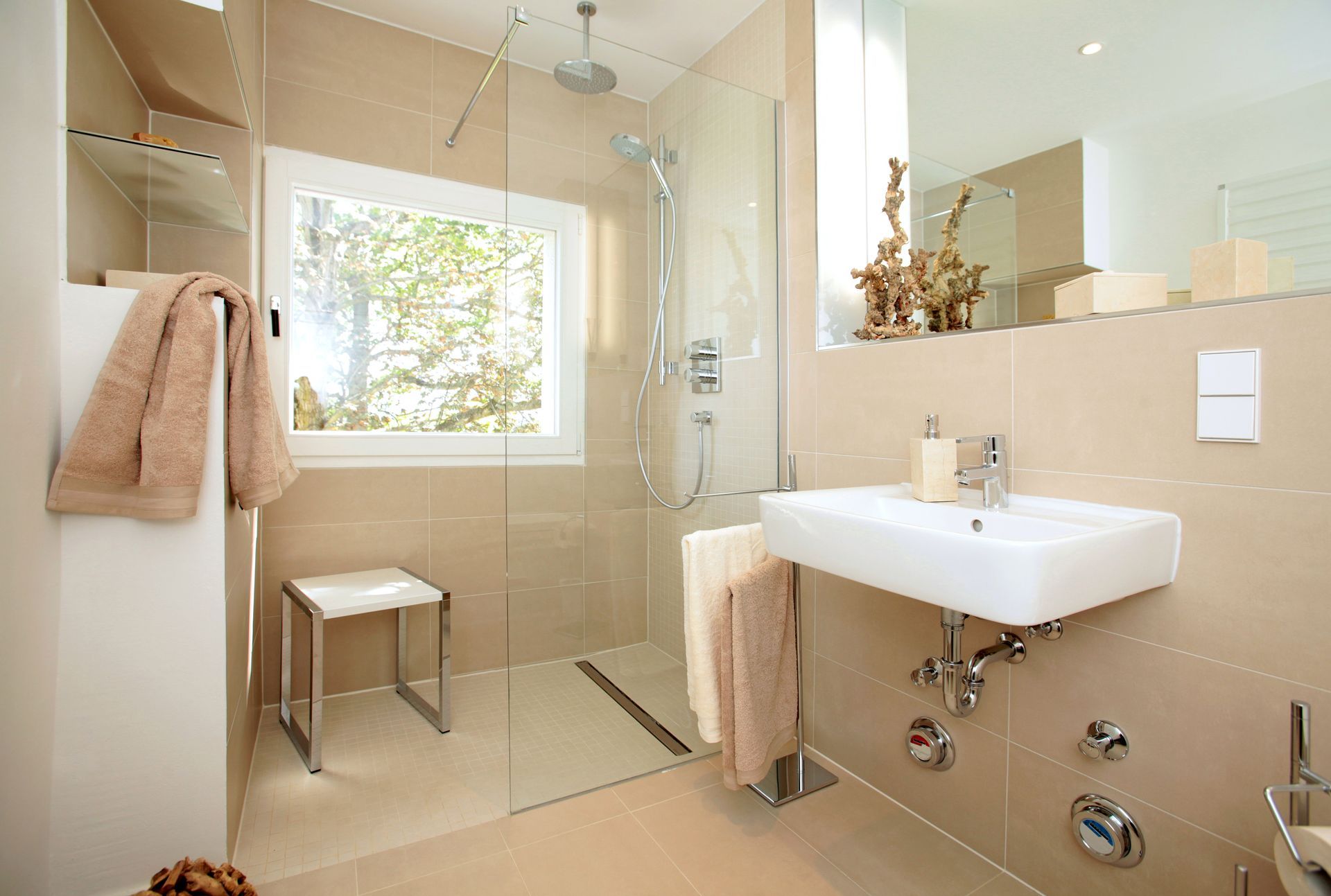 Modern bathroom with shower, sink, and window. Beige tiles, glass shower screen, and a stool.