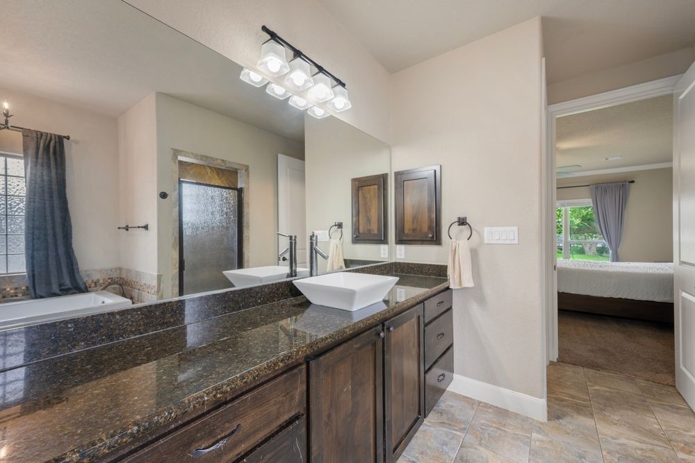 Bathroom with brown vanity, white sink, large mirror, and open doorway to bedroom.