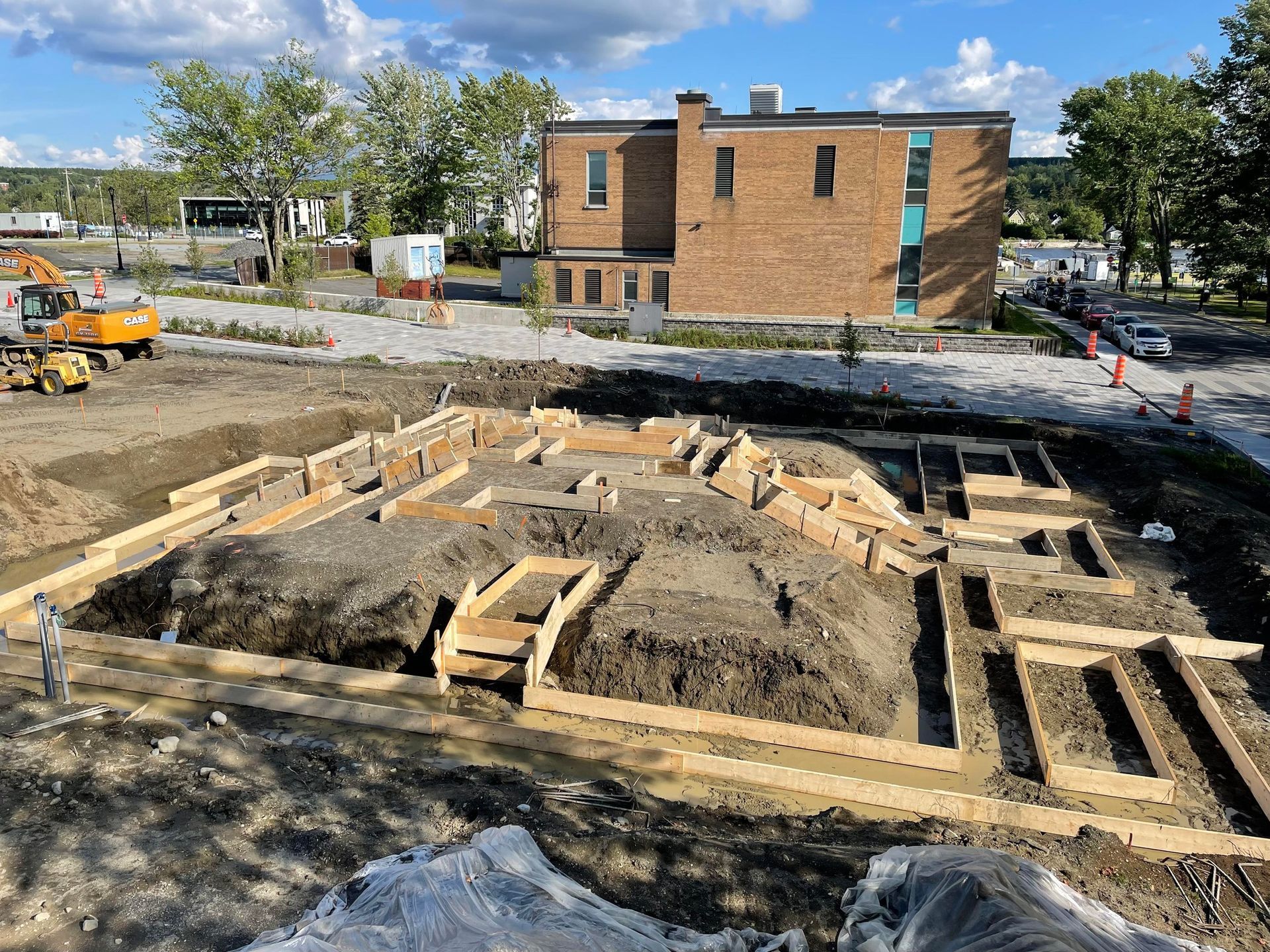 Chantier de construction avec coffrages en bois pour les fondations ; bâtiment en briques en arrière-plan.