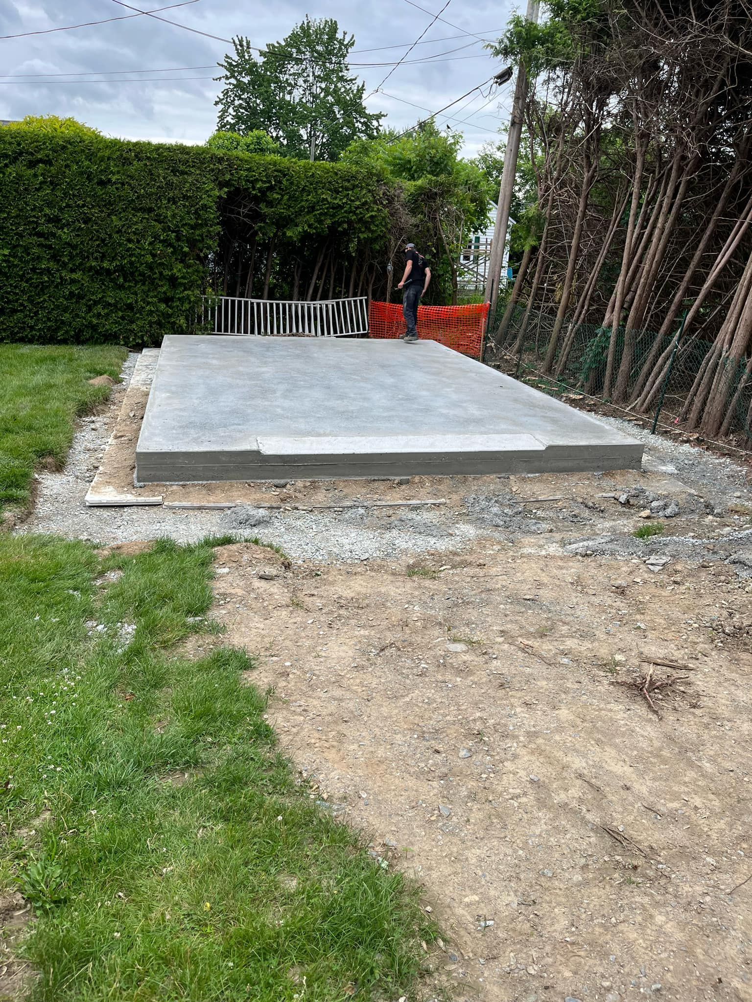 Fondation en béton entourée de gravier, gazon et une personne dans un jardin, avec une haie en arrière-plan.